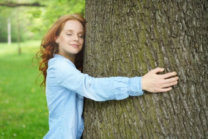 Eine junge Frau mit langen roten Haaren umarmt einen Baum auf einer Wiese und lächelt dabei.