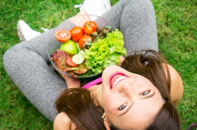 Eine Frau sitzt im Schneidersitz auf einer Wiese, in ihrem Schoß befindet sich eine große Schale mit Salat. Sie blickt nach oben in die Kamera und wirkt dabei sehr zufrieden.