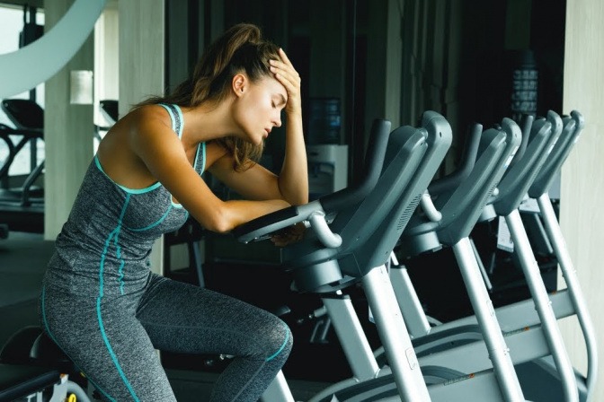 Eine junge Frau in kompletter Sportmontur sitzt auf einem stationären Fahrrad im Fitnessstudio. Sie hat den Kopf auf dem angewinkelten rechten Arm gestützt und sieht verzweifelt und gestresst aus.