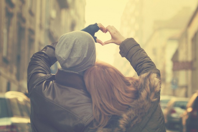 Ein Paar spaziert durch eine Straße und formt gemeinsam mit der jeweils linken und rechten Hand ein Herz. 