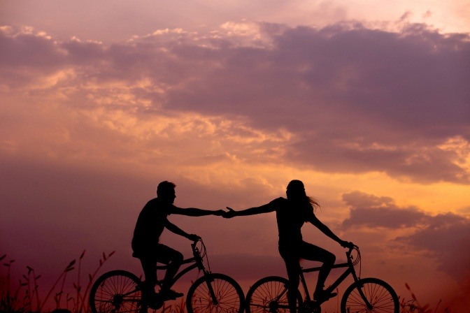 Ein Pärchen fährt gemeinsam mit dem Fahrrad einen Berg hoch, während hinter ihnen die Sonne untergeht. 