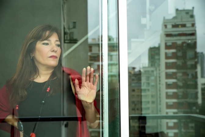 Reife Frau, die in einer urbanen Umgebung aus dem Fenster ihrer Wohnung schaut und nachdenklich wirkt.