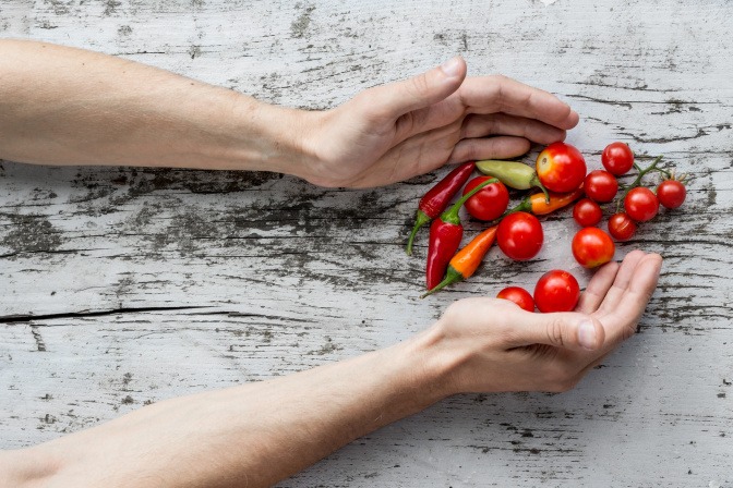 Einige Tomaten und rote Paprika liegen auf einer grauen Holzoberfläche. Jemand formt mit den Händen eine Schale, um sie aufzugreifen.