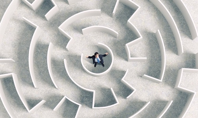 Das Bild zeigt einen Blick aus der Vogelperspektive auf ein Labyrinth. Im Zentrum steht ein einsamer Mann und blickt fragend nach oben.