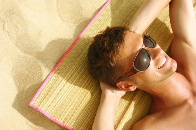 Ein Mann mit einer Sonnenbrille liegt auf einer Matte am Strand und lächelt