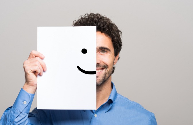 Ein glücklich wirkender und lächelnder Mann hält eine Zeichnung mit einem Smiley vor die linke Seite seines Gesichts.