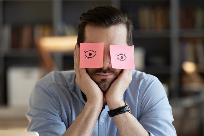 Mann zeigt keine Gefühle - ein Mann sitzt apathisch am Tisch, über seinen Augen kleben rosafarbene Post-its, auf denen Augen aufgemalt sind.
