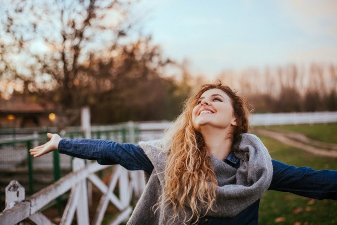 Eine junge Frau seht auf einer Weide und breitet die Arme aus, sie hat einen glücklichen und zufriedenen Gesichtsausdruck.