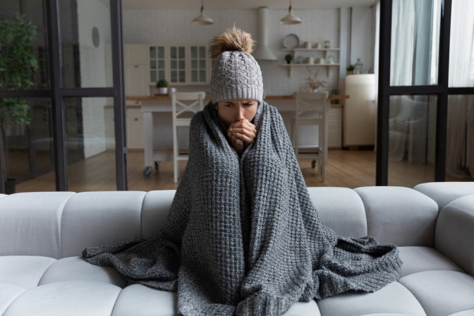 Frau sitzt mit Decke und Mütze auf dem Sofa