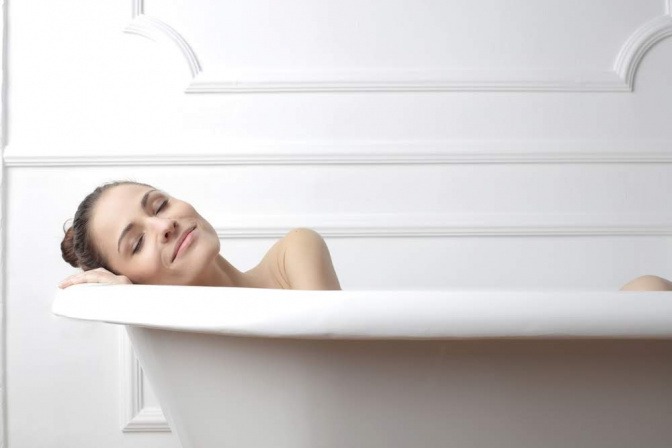 Eine Frau liegt in der Badewanne. Sie hat die Augen geschlossen, ihr Kopf liegt auf dem Seitenrand und sie wirkt zufrieden und entspannt.