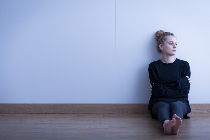 Traurig und nachdenklich wirkende junge Frau sitzt in dunklem Raum auf Boden und lehnt an Wand.