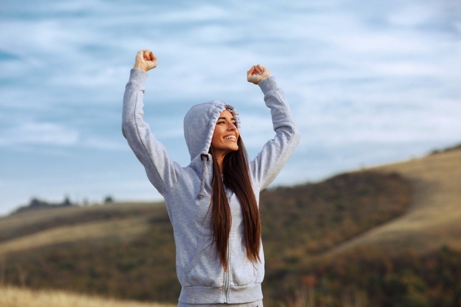 Eine junge Frau in der Natur hebt motiviert ihre Hände und lächelt