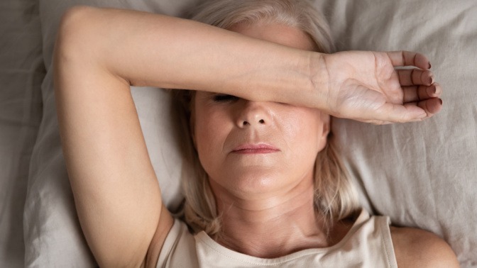 Müde Frau in den Wechseljahren liegt im Bett und hält den Arm über ihr Gesicht