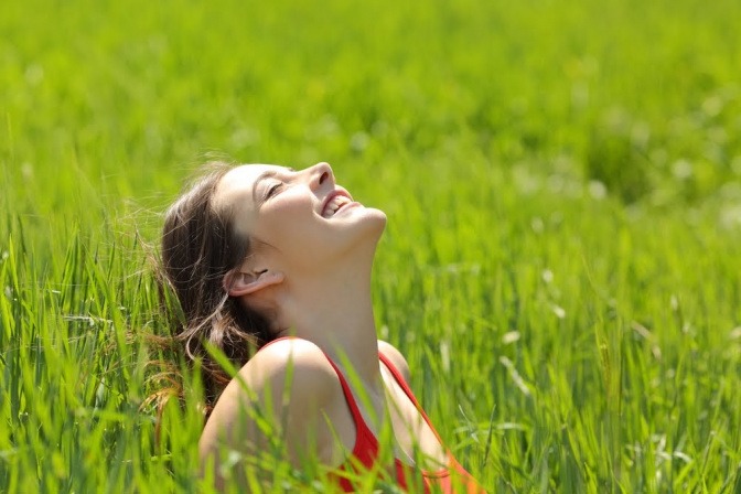 Eine Frau sitzt auf einer Wiese mit hohem Gras und lächelt gen Himmel.