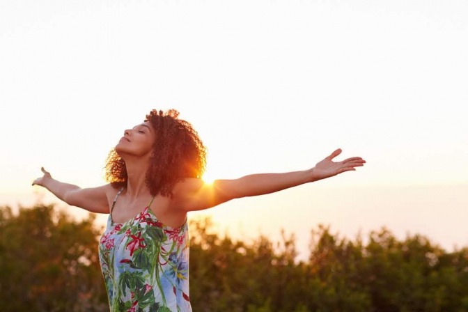 Eine Frau steht beim Sonnenaufgang mit ausgebreiteten Armen auf einem Feld und atmet tief ein.