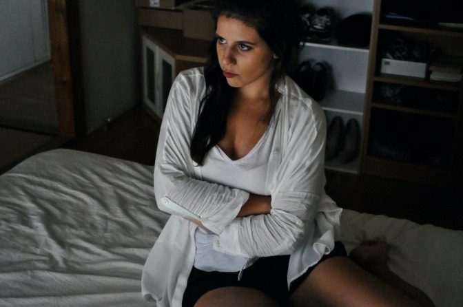 Eine Frau in einem weißen Cardigan sitzt auf einem Bett. Sie wirkt frustriert.