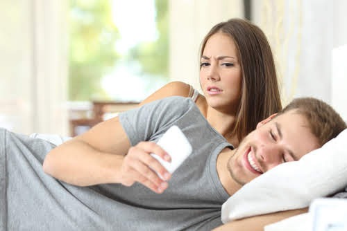 Ein Paar liegt im Bett, er liest eine Nachricht auf seinem Handy und lächelt, seine Frau blickt skeptisch und verunsichert in seine Richtung.