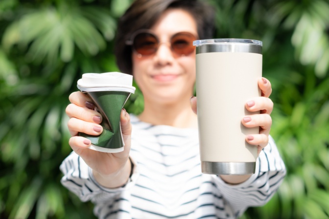 Frau hält in der einen Hand einen zusammengedrückten Kaffeebecher aus Plastik und in der anderen einen wiederverwendbaren Coffee-to-go-Becher.   