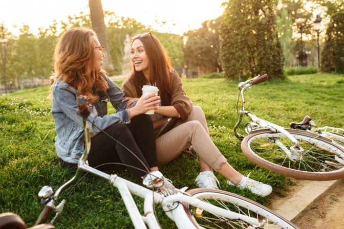 Zwei junge Frauen mit Fahrrädern, sitzen als Freunde auf einer Wiese in einem Park.