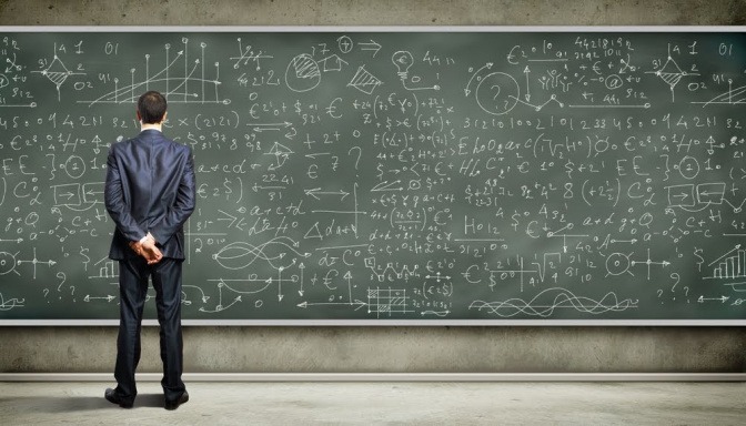 Ein Mann in einem Anzug steht vor einer großen Tafel, die vollgeschrieben ist mit komplizierten Formeln und Gleichungen.