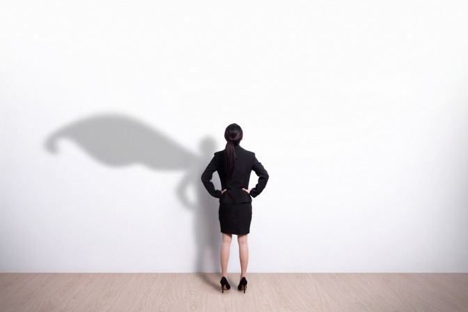 Eine Business-Frau steht vor einer weißen Wand, an der sich ihr Schatten in Form eines Superhelden abzeichnet