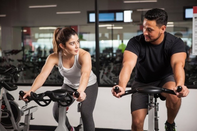 Ein junger Mann und eine junge Frau sitzen im Fitness-Studio je auf einem Radtrainer und unterhalten sich während des Trainings.