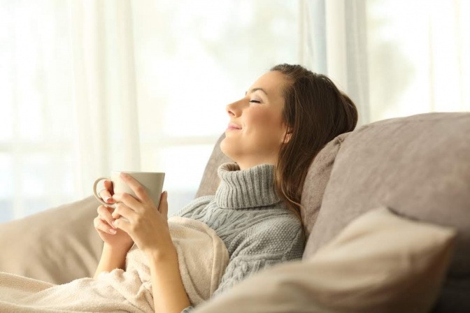 Eine Frau liegt auf dem Sofa und hält eine Tasse in den Händen. Ihr Kopf lehnt an der Rückenlehne, die Augen sind entschlossen. Sie wirkt entspannt.