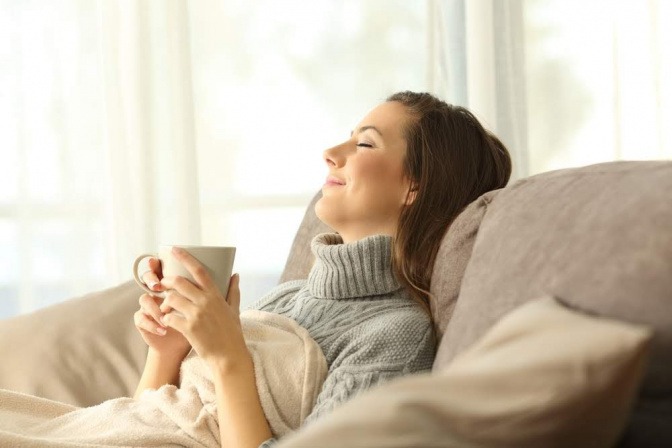 Eine Frau liegt auf dem Sofa und hält eine Tasse in den Händen. Ihr Kopf lehnt an der Rückenlehne, die Augen sind entschlossen. Sie wirkt entspannt.