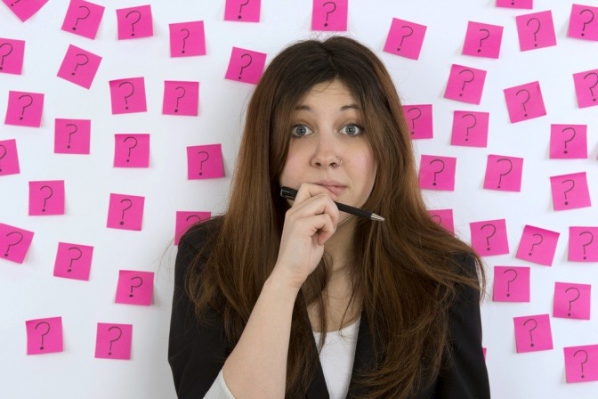 Frau steht mit fragendem Gesicht vor einer Wand, auf der viele Notizzettel mit Fragezeichen kleben.