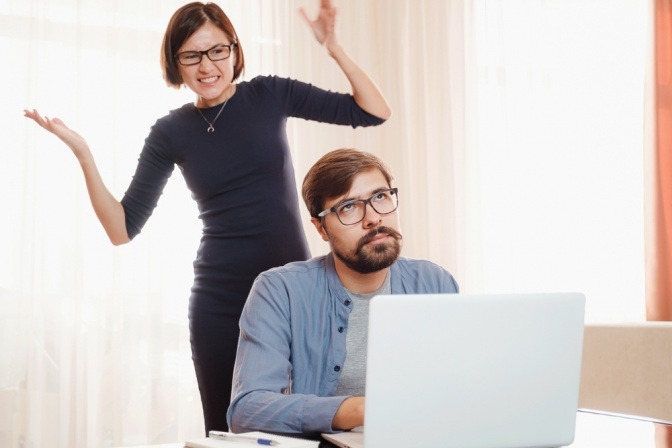 Paar streitet sich, er sitzt mit genervtem Ausdruck am Laptop, seine Partnerin steht verärgert und gestikulierend hinter ihm.