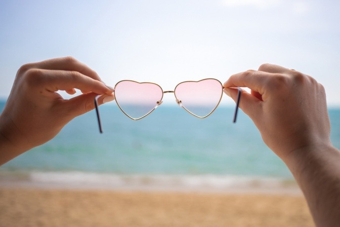 Eine rosarote Brille in Herzform wird am Strand von zwei Frauenhänden gehalten 