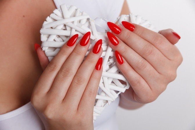 Frauenhände mit roten Fingernägeln halten ein herzförmiges, weißes Objekt fest. 