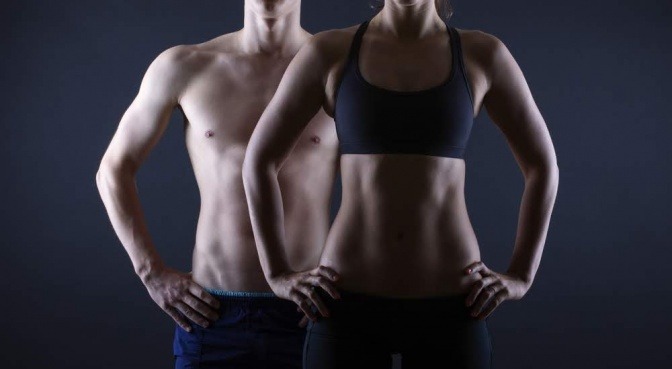 Der Bildausschnitt zeigt die Oberkörper eines Mannes und einer Frau. Sie stehen leicht versetzt nebeneinander, er mit freiem Oberkörper, sie trägt einen Sport-BH. Beide wirken sehr fit. Der Fokus des Bildes liegt auf der Körpermitte. 