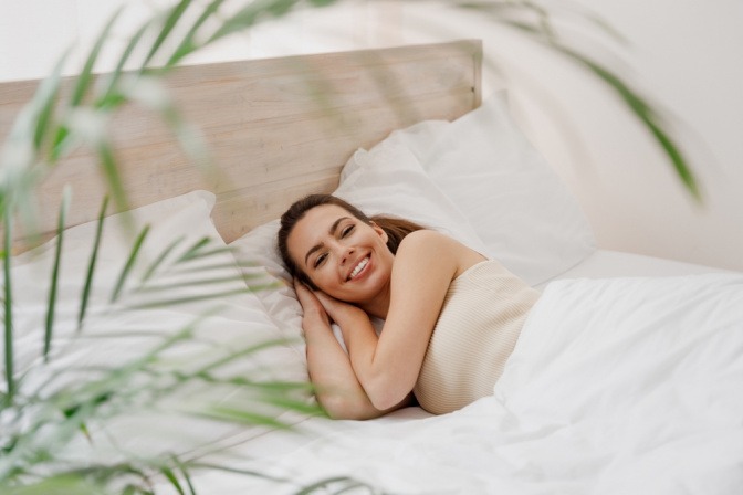 Zufriedene und erholt wirkende Frau wacht in ihrem Bett in einem Schlafzimmer mit Pflanzen auf.