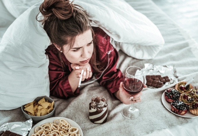 Eine Frau liegt im Bett, trinkt Wein, raucht und will schlechte Gewohnheiten loswerden.