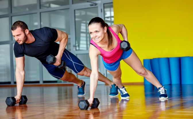 Eine Frau und ein Mann machen Sport. Sie befinden sich in einem Raum mit gelben Wänden. Die Übung ist ähnlich wie eine Liegestütze, bloß haben die Personen Handeln in den Händen.