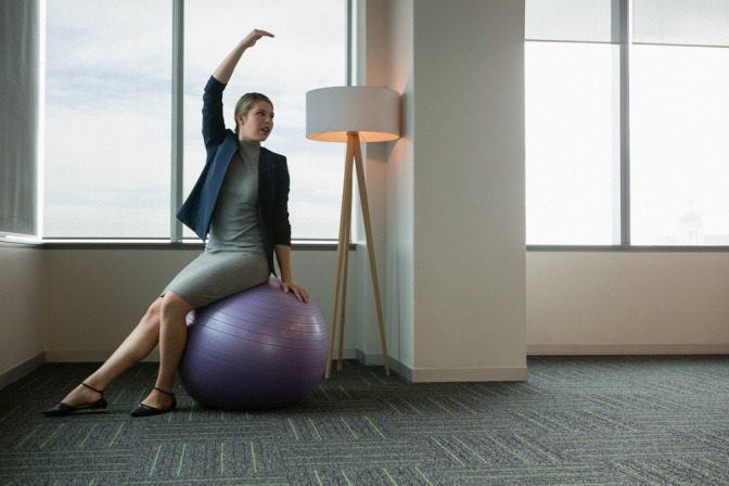 Eine Frau im Businessoutfit macht Übungen auf einem Gymnastikball.