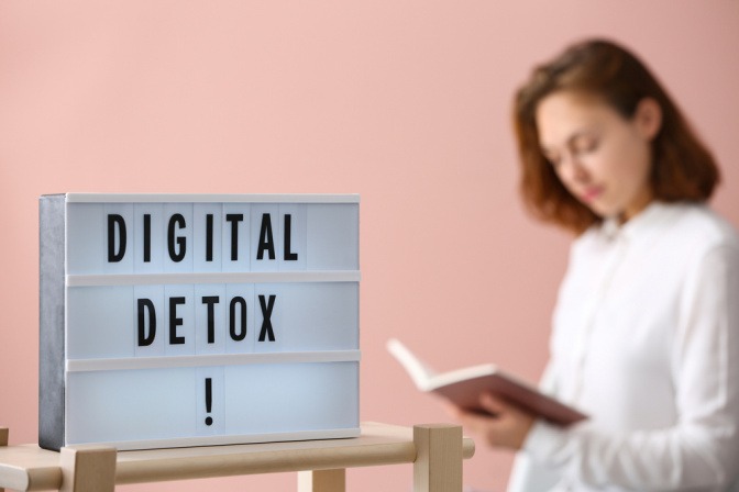 Leuchtreklame mit der Aufschrift "Digital Detox". Im Hintergrund liest eine Frau ein Buch.