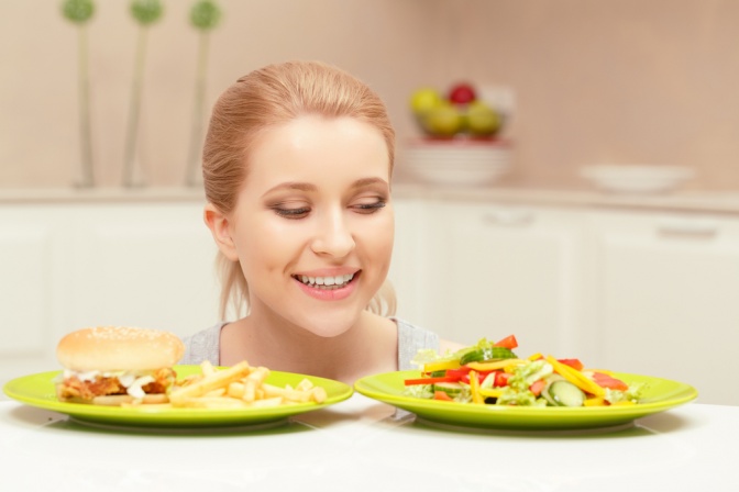Auf dem Foto sind zwei Teller und eine blonde Frau abgebildet. Auf dem einen Teller befindet sich gesundes, auf dem anderen ungesundes Essen.