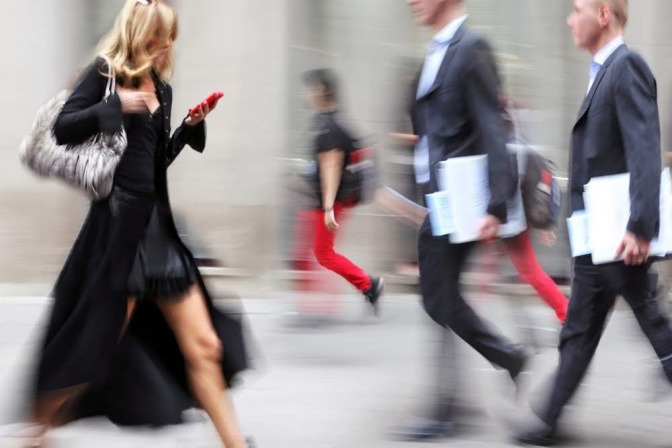 Eine elegant gekleidete Frau schreitet durch eine belebte Innenstadt. Alles wirkt hektisch, sie starrt beim Laufen auf ihr Smartphone und könnte Tipps für mehr Bewusstsein sicherlich gut gebrauchen.
