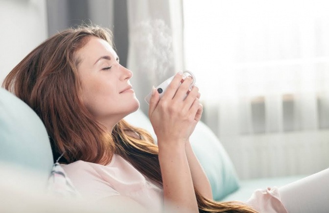 Eine Frau sitzt mit geschlossenen Augen auf einem Sofa, hält eine Tasse Tee in der Hand und wirkt sehr entspannt.