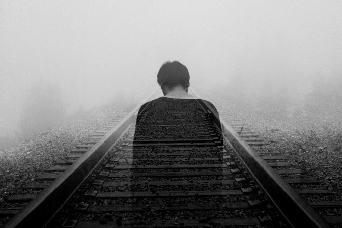 Ein Mann steht im Nebel und bei Regen am Anfang einer endlos wirkenden Gleisstrecke.