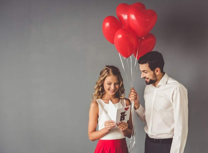 Ein Mann macht einer Frau während eines Dates ein Geschenk, das aus mehreren roten Luftballons und einer romantischen Karte besteht.