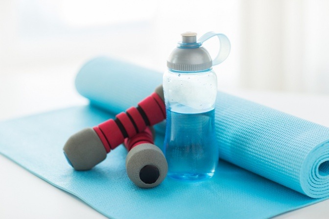 Eine Trinkflasche steht auf einer Sportmatte, daneben liegt eine Hantel
