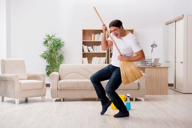 Ein junger Mann räumt mit viel Enthusiasmus seine Wohnung auf und funktioniert einen Besen dabei zur Luftgitarre um.