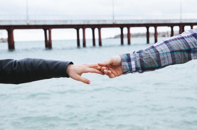 Mann und Frau halten an einem Strand Händchen, die Verbindung droht sich jedoch aufzulösen, beide sind nur noch an Fingerspitzen verbunden.