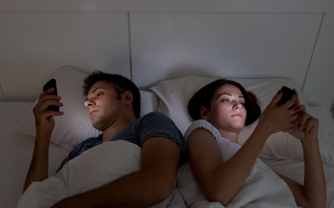 Junges Paar liegt nebeneinander im Bett, beide starren auf ihre Smartphones und kümmern sich nicht um den Partner.