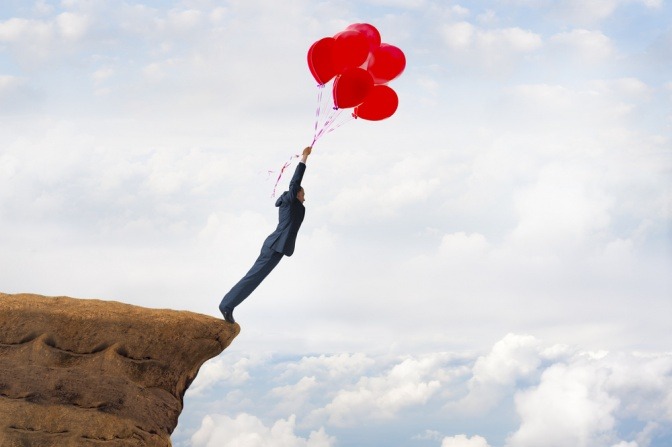 Mann hängt an Luftballons und springt mutig von einem Felsvorsprung.