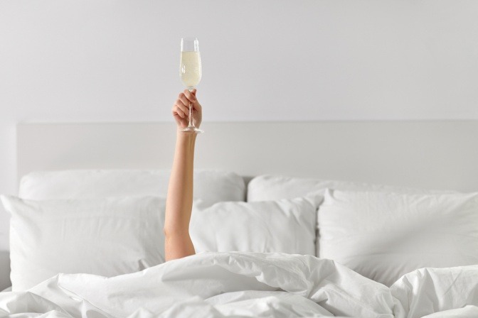 Eine Frau liegt unter Bettdecken begraben im Bett und hält ein gefülltes Sektglas hoch. 