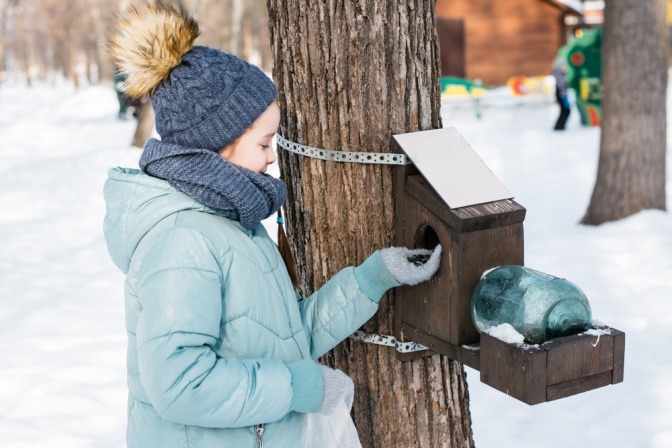 Mädchen füllt in schneebedecktem Garten Futterhäuschen für Vögel nach.